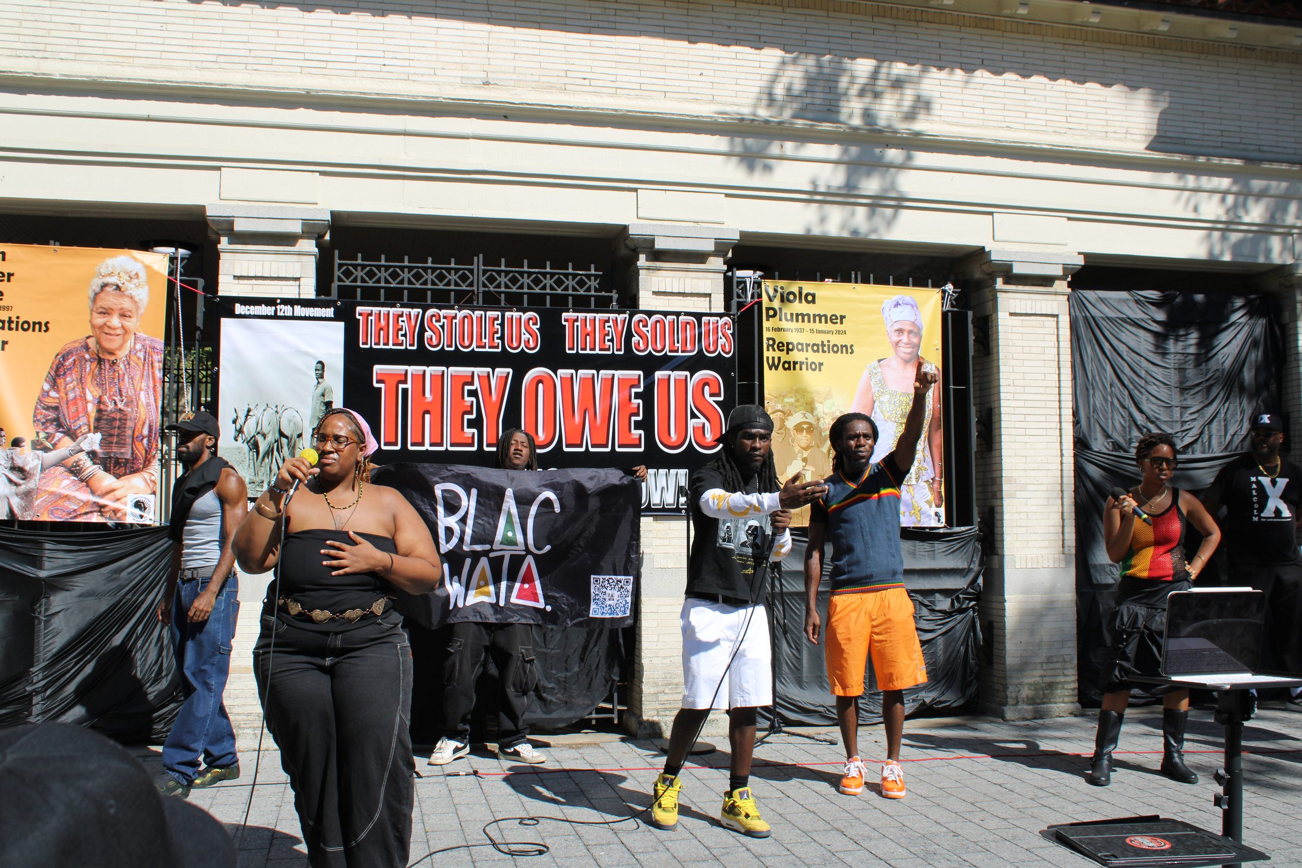 Speakers on stage in front of a "They Stole Us, They Sold Us, They Owe Us" banner